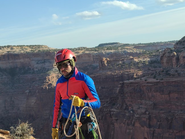 lt= park Ranger with climbing rope and harness stands near edge of canyon.