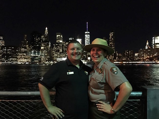 Nighttime scene of a man in blue shirt with park ranger spouse with New York City in background.