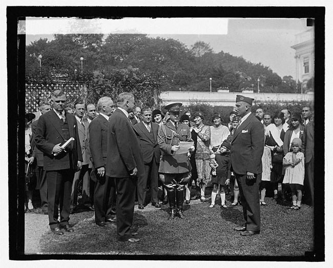 A crowd of men in suits and military uniforms, surrounded by an audience of women and children A crowd of men in suits and military uniforms, surrounded by an audience of women and children