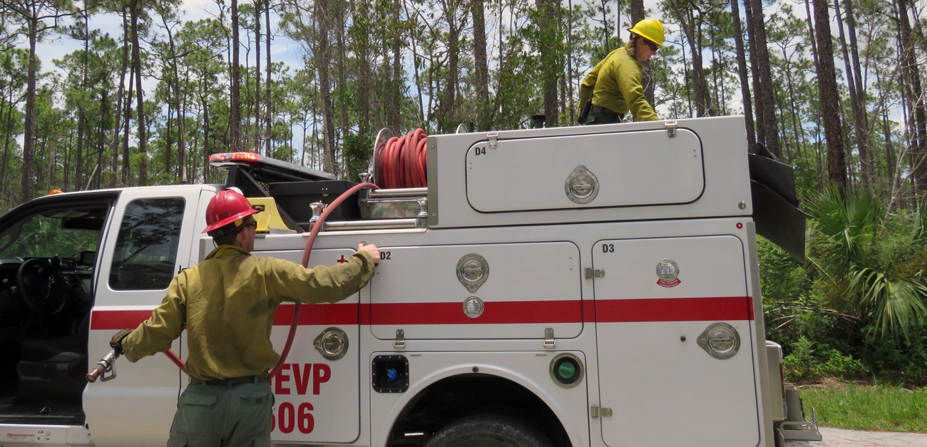 A firefighter pulls a hose down from an engine while another firefighter on top of the engine leans over.