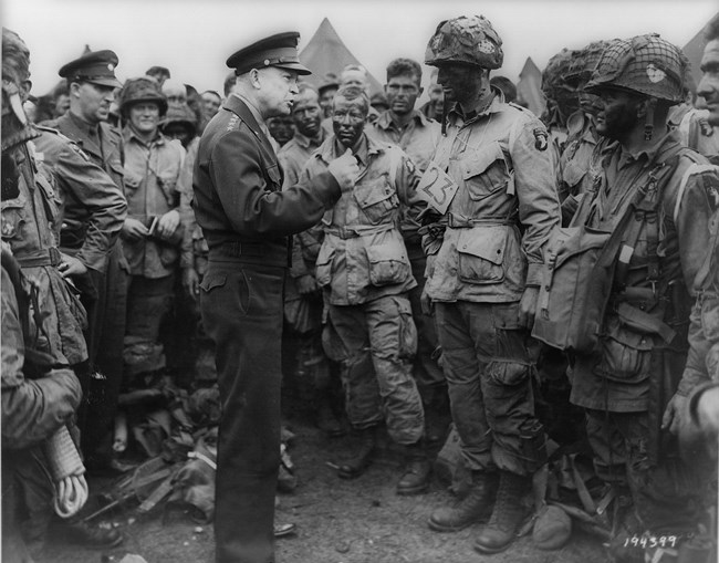 A black and white photo shows General Dwight D. Eisenhower speaking to a group of soldiers. Eisenhower stands in the middle of the photo, wearing a neat, dark uniform, a billed cap, and four stars on his shoulder. The soldiers face Eisenhower. Their faces
