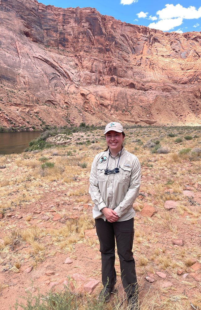 Person stands on grassy sandy shoreline near rivers edge at the bottom of a tall red canyon.