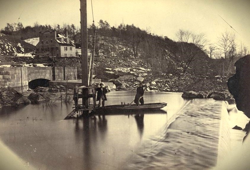 Sepia tone image showing two men standing in a small flat boat, on water, near the spillway of a dam. The men wear hats, jackets, and trousers. Behind them is a snowy hill with bare trees and stone wall and stone building