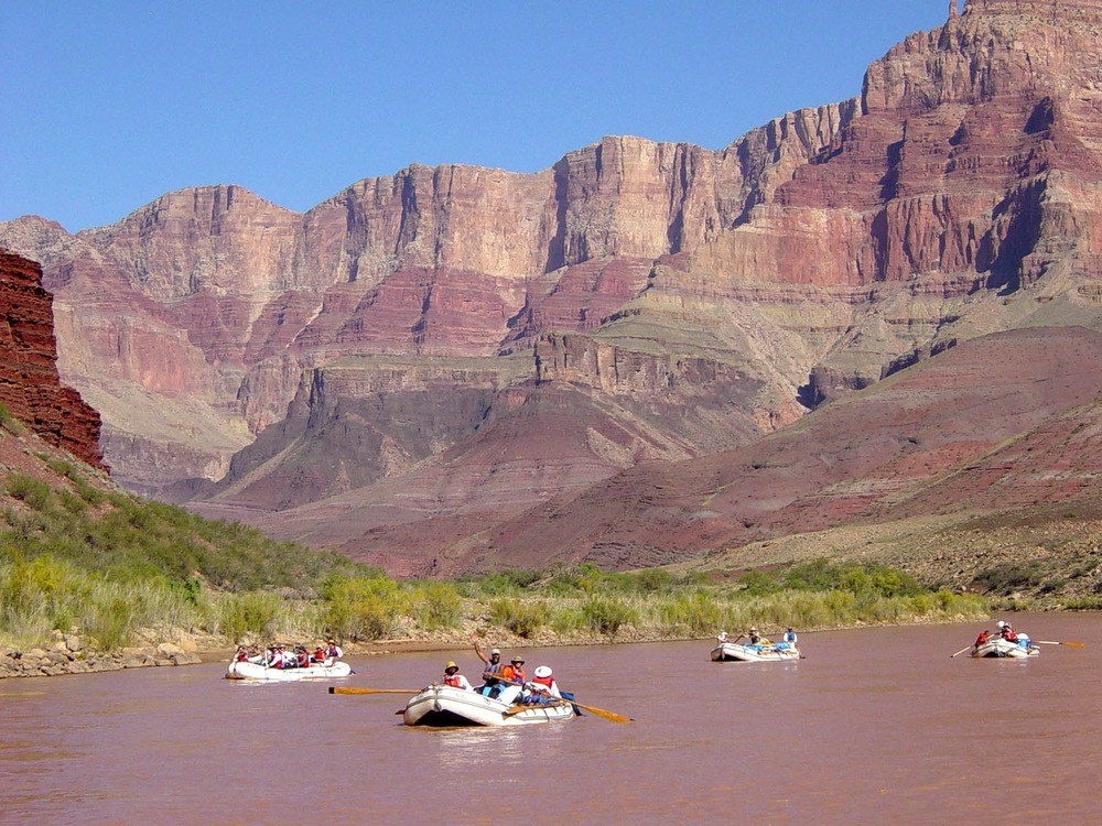 4 rafts floating down river with canyon wall in background.