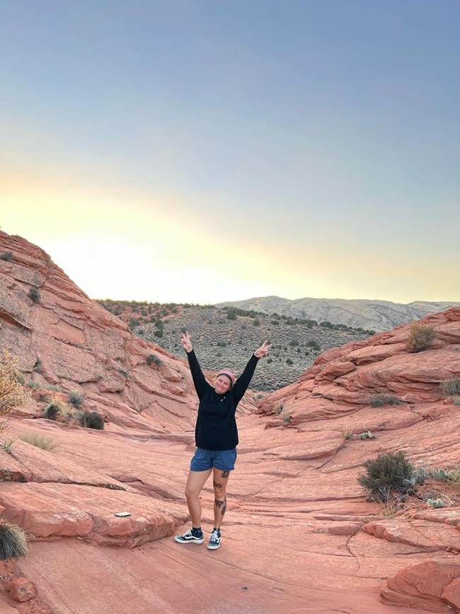 Kelsey Shores Park Story 2 A person holds up two peace signs with their fingers and poses near a sloping red rocks.