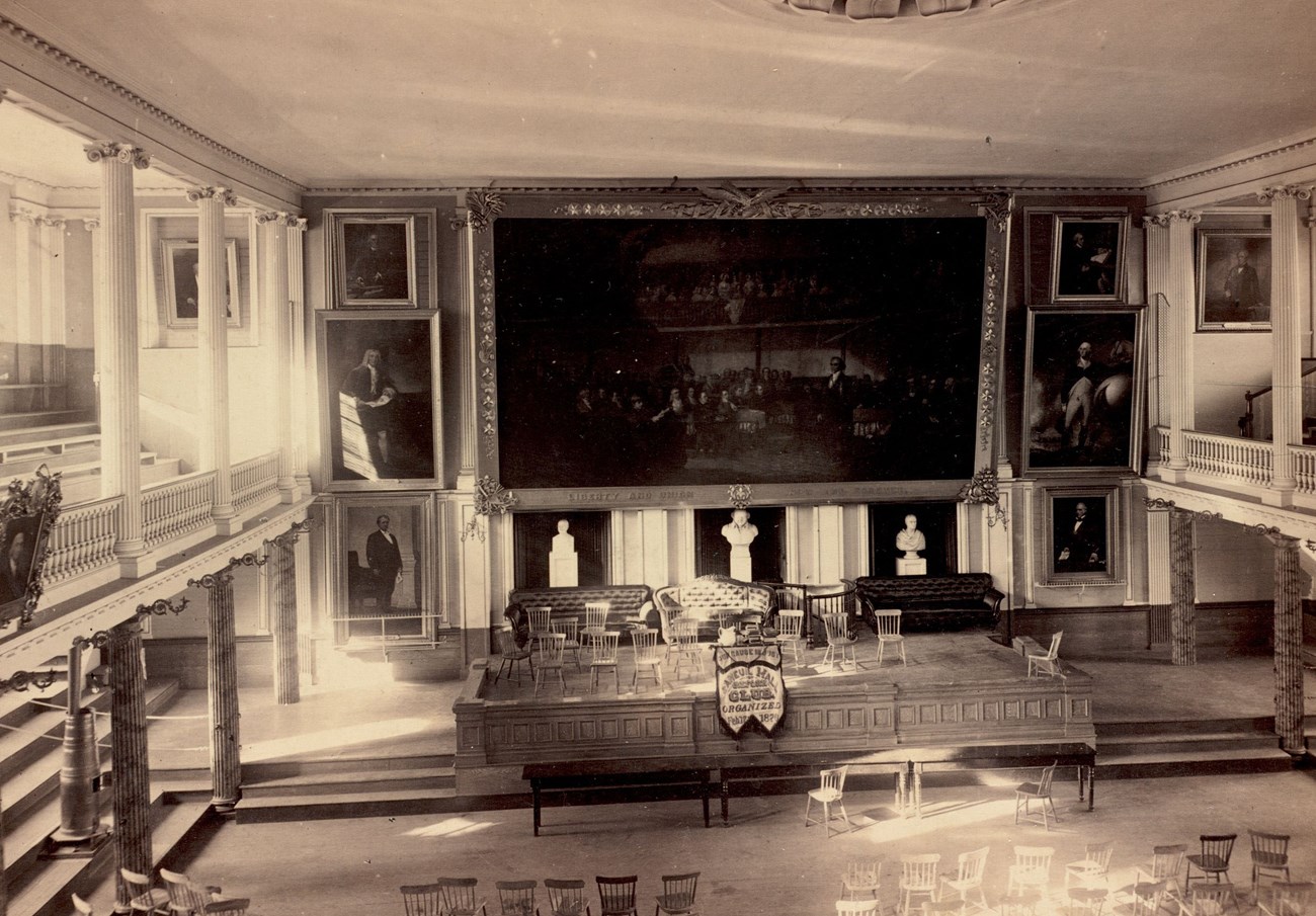 FHinteriorBPL view from gallery of Faneuil Hall, an open hall with a stage and large paintings on the walls.