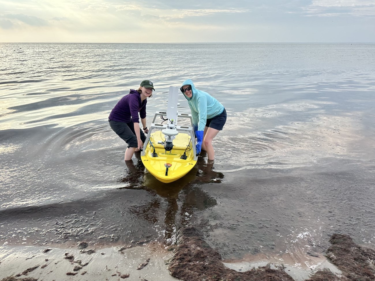 Two people squat in water that is ankle-deep and smile while holding a small yellow boat in the water.