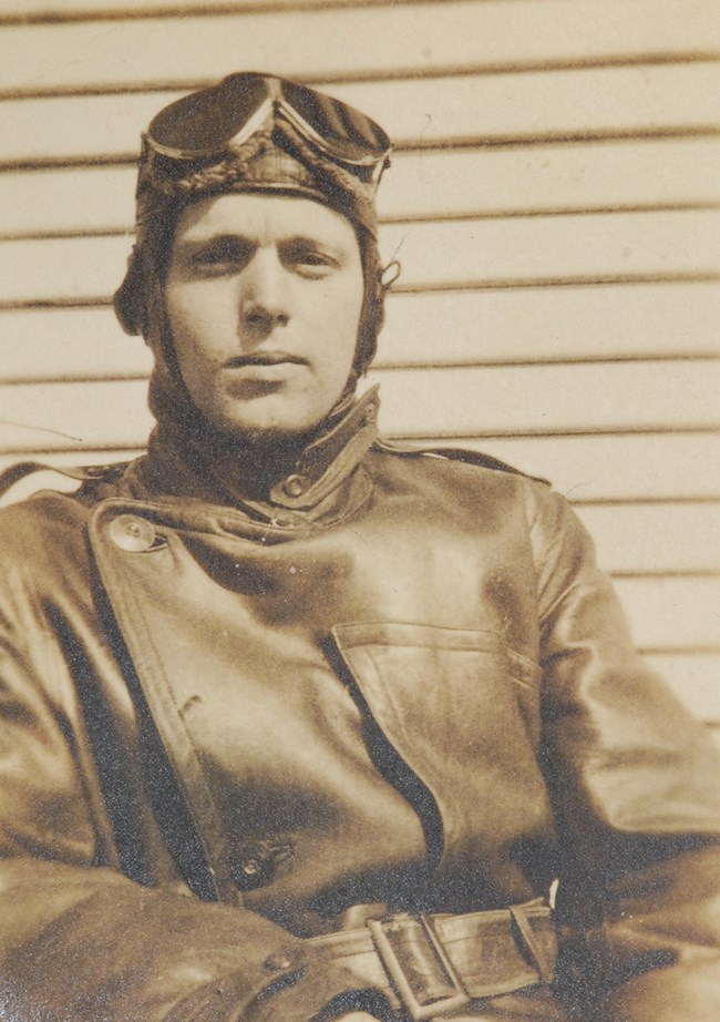 Black and white photo of man wearing leather jacket, cowl, and flight goggles.