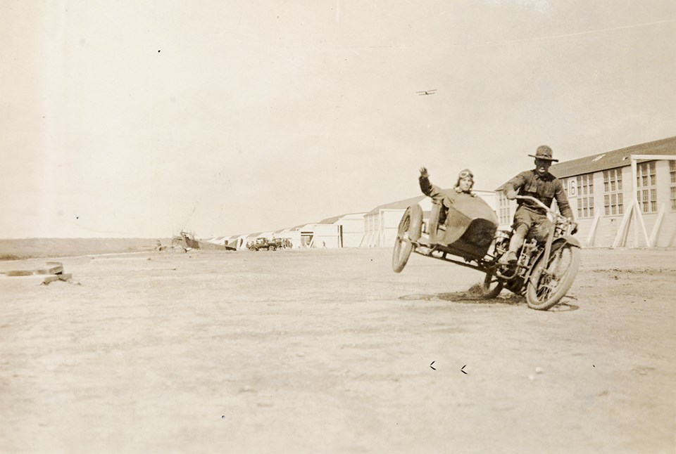 Black and white photo of man riding a motorcycle with another man in sidecar in front of airplane hangars.