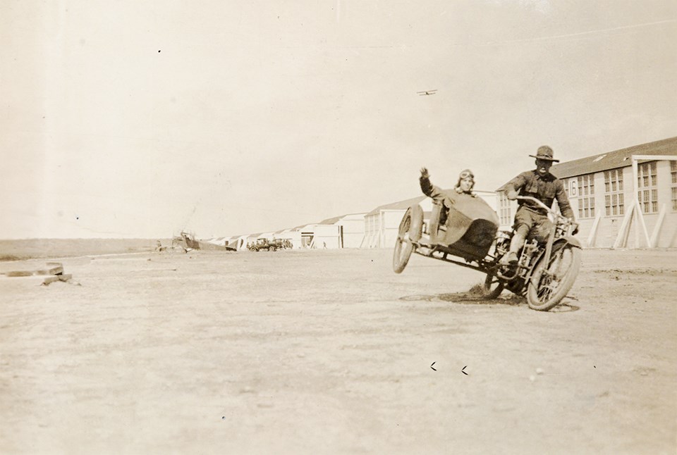 Jazzin a little while Black and white photo of man riding a motorcycle with another man in sidecar in front of airplane hangars.