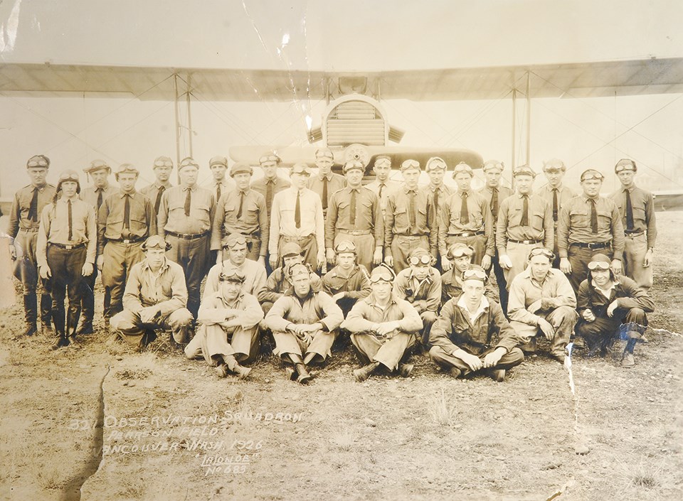 Group of men wearing pilot's goggles and suits pose in front of biplane aircraft.