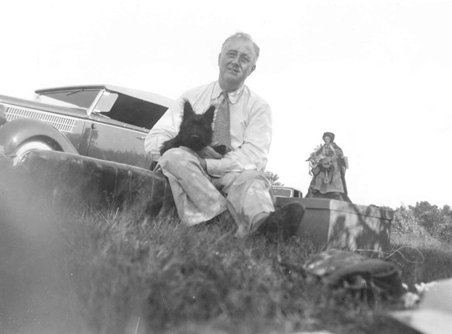 Man in white trousers, white shirt, and tie sits near the ground. He has a black Scottish Terrier dog in his lap. A classic car and a statue are in the background.
