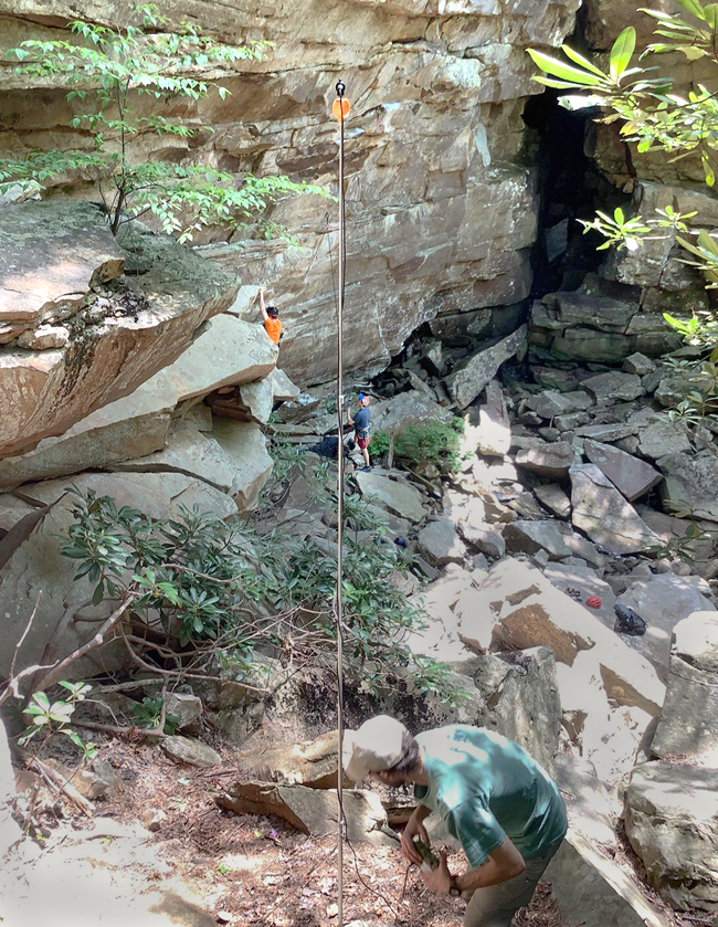 Acoustic detector A person is next to a long pole with an orange circle at the top which is set up next to a cliff face where there are two people climbing.