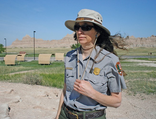 A woman stands in park ranger uniform with blue skies and buttes in the background.