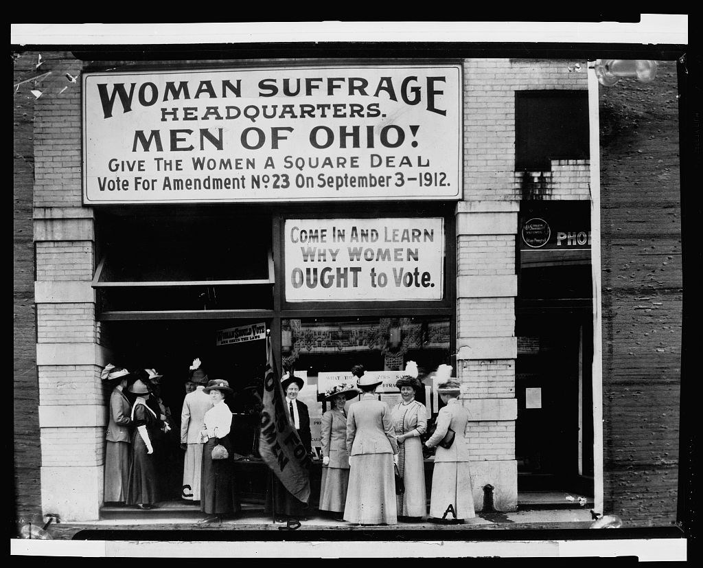 A black and white photo of women in early 1900s attire standing in front of a brick building bearing a sign: "Woman Suffrage Headquarters. Men of Ohio! Give the Women a Square Deal".
