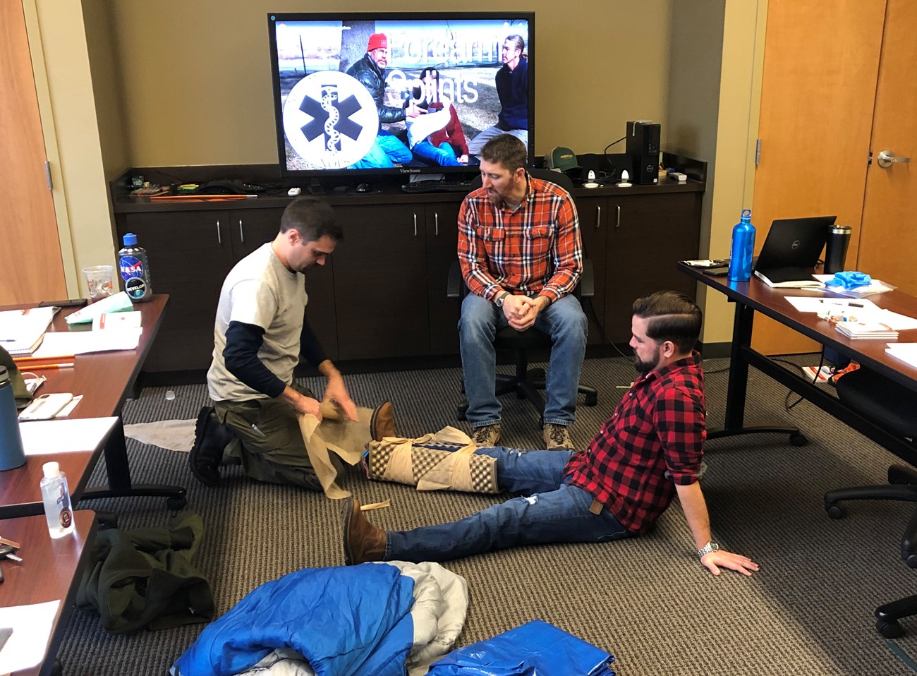 Man places a splint on a another man's leg while third man watches.
