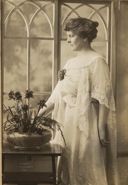 Formal portrait, three-quarter-length, Florence Brooks Whitehouse, facing left, standing by window and table with container of flowers, wearing loosely flowing dress with corsage on right lapel.
