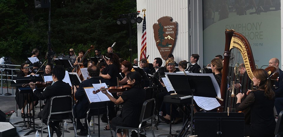 A symphony performs outdoors in a white amphitheater. The projector screen behind them reads, "Symphony at the Seashore."