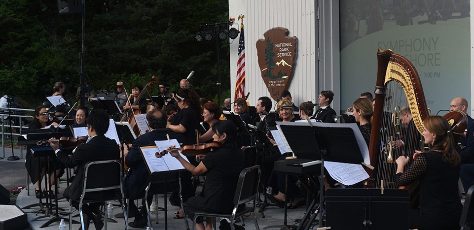 The Cape Symphony performs at the Salt Pond Visitor Center amphitheater. A symphony performs outdoors in a white amphitheater. The projector screen behind them reads, "Symphony at the Seashore."