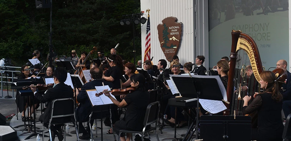 The Cape Symphony performs at the Salt Pond Visitor Center amphitheater. A symphony performs outdoors in a white amphitheater. The projector screen behind them reads, "Symphony at the Seashore."