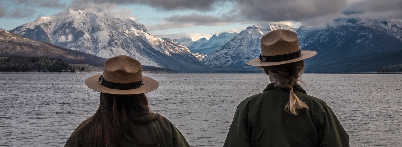 GLAC McDonald Lake Back of two rangers in flat hats looking a lake in front of a mountain