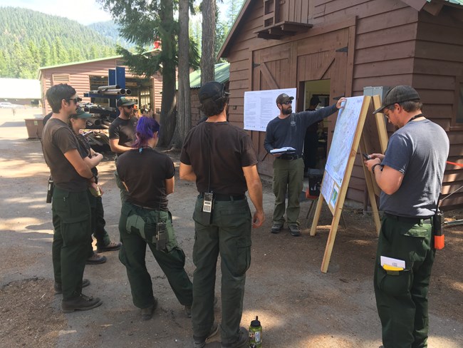 A group of six individuals listen to another individual next to a map.