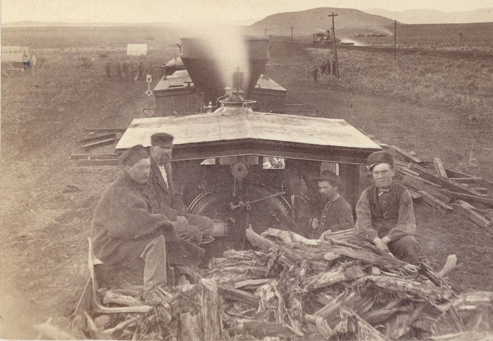 Black and white photo of railroad workers sitting on top of wood piled in a train cart