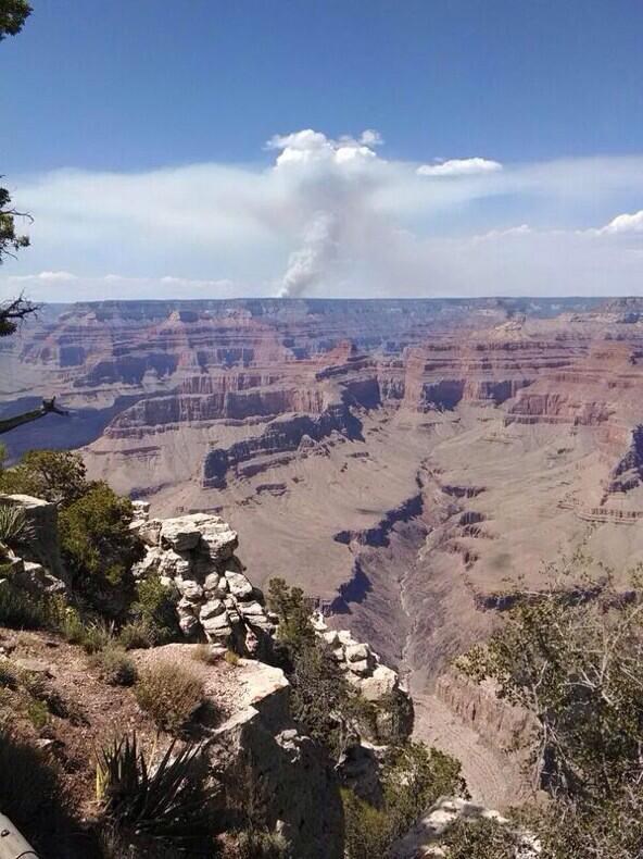 A plume of smoke in the distance beyond the Grand Canyon rises into the blue sky.