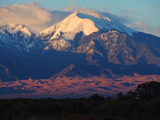 large snow-capped mountain with forested slopes and sand dunes in the foreground