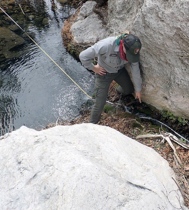 A person straddling a measuring tape and flow of water off of an opening between boulders into a natural pool.