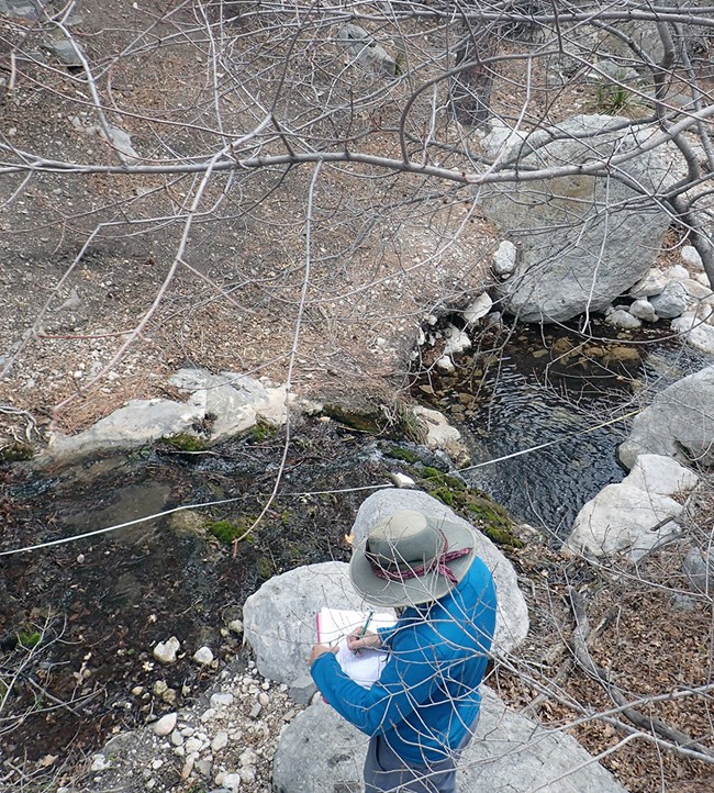 A person writing on a clipboard standing on a rock overlooking a channel of water with a measuring tape stretched across it