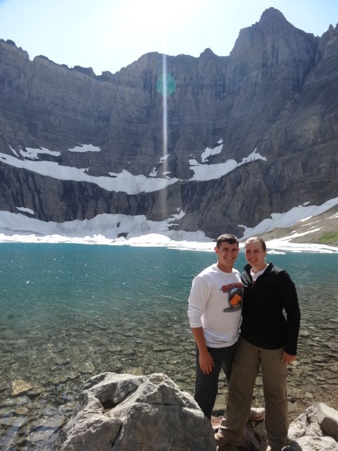 A man and woman standing by a glacial lake with mountains in the background.