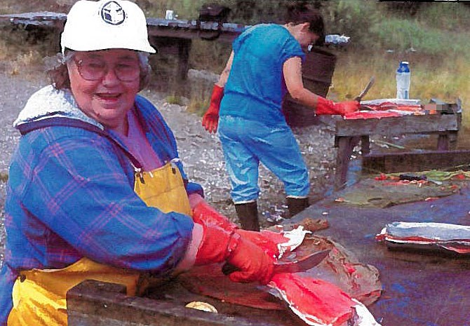 Cutting Salmon An older woman wearing a white ball-cap, blue jacket and waterproof, yellow apron cutting fish on a wet wooden table.