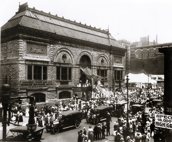 Women in white on building steps with numerous onlookers standing around