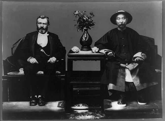 two men sitting in chairs with a table, vase, and flowers between them.