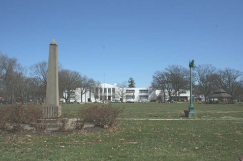 Tall, grey, stone monument on green grass and white building in the background.