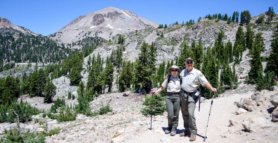 Greg and Annette Brahms on the Bumpass Hell Trail in Lassen Volcanic National Park. A man and a woman on a rocky trail backed by a large peak.