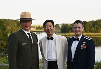 Three men stand outdoors in front of a large pond ringed with trees. From left to right: A man wearing a green National Park Service dress uniform, a man in a tuxedo with a white coat, and a man wearing a blue United States Air Force dress uniform.