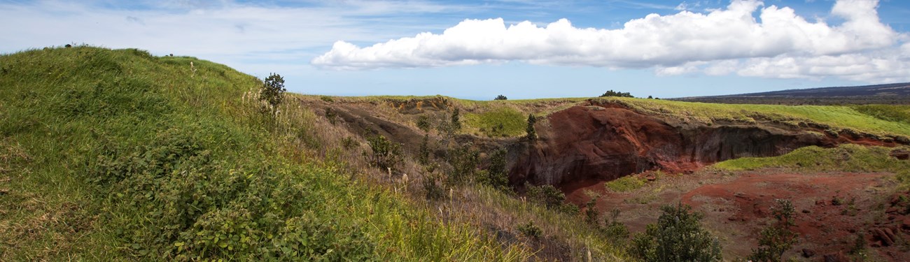 Green hill with red volcanic soil exposed under blue sky with clouds