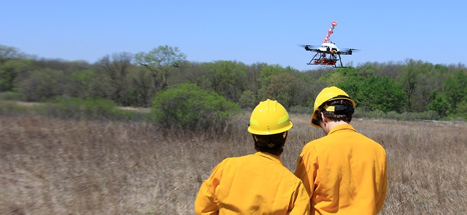 An unmanned aircraft hovers above a field while two men in firefighting gear stand nearby.