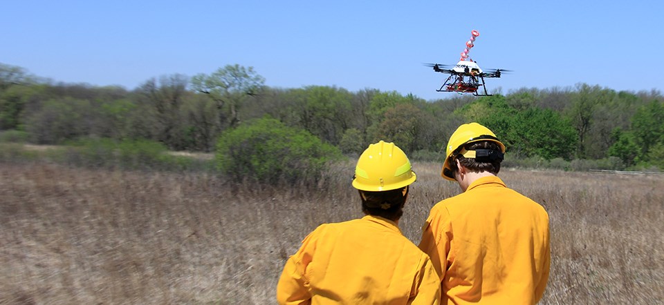 An unmanned aircraft hovers above a field while two men in firefighting gear stand nearby.