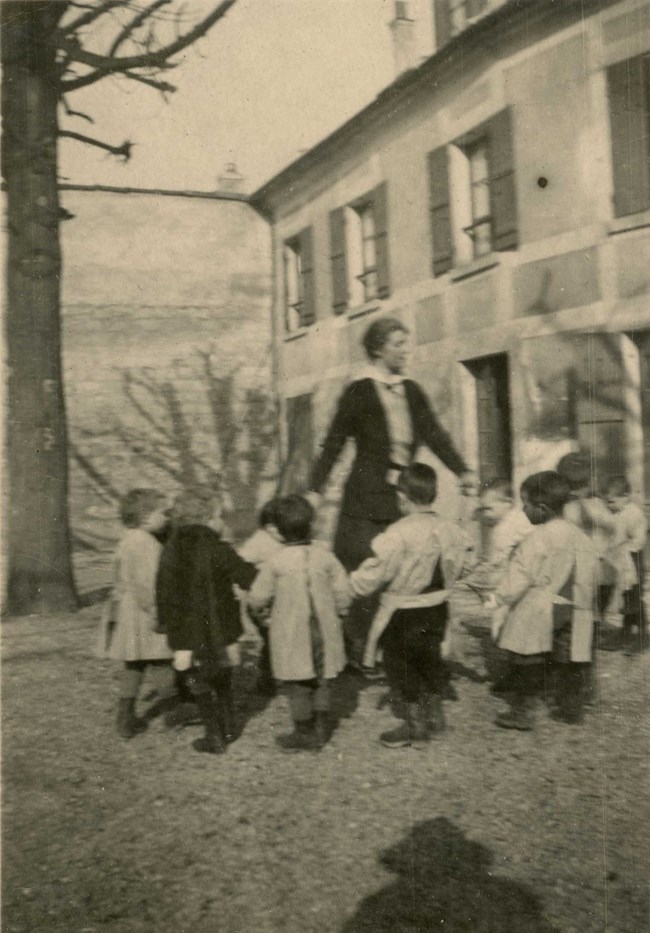 Woman and about nine small boys hold hands in a ring in front of building.