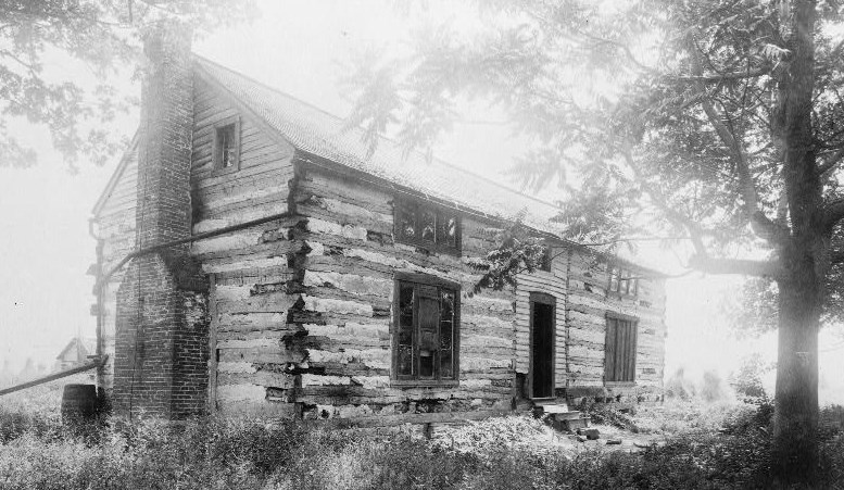 Black and white photo of a two-story, four-room log cabin surrounded by trees