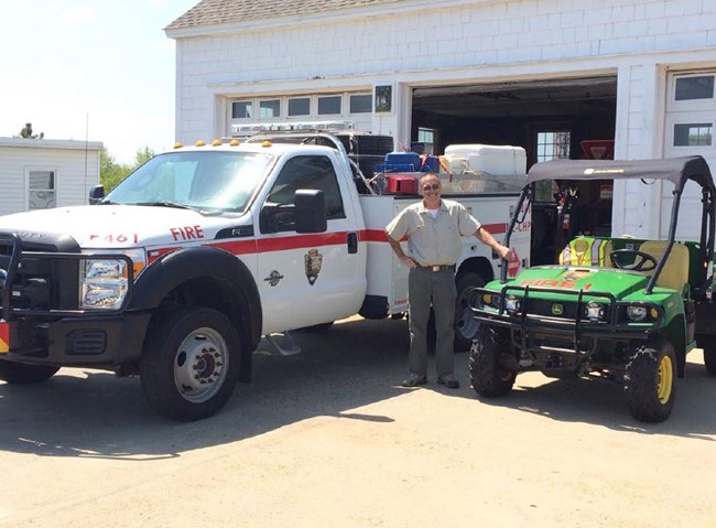 Man standing in front of a white truck that says "fire" and next to a all terrain vehicle.