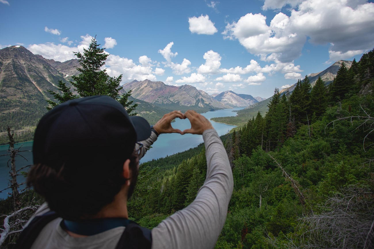 A hiker holds up their hands in the shape of a heart. Mountains and lake in background.