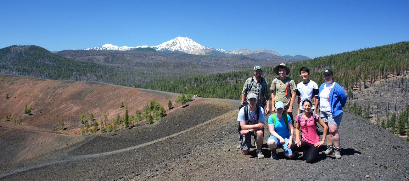 A group of seven people pose for a photo on the crater of a cinder cone volcano; a snow-capped volcano rises in the distance