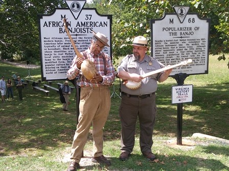 Two men hold banjo-like instruments. One man wears a Park Service uniform.