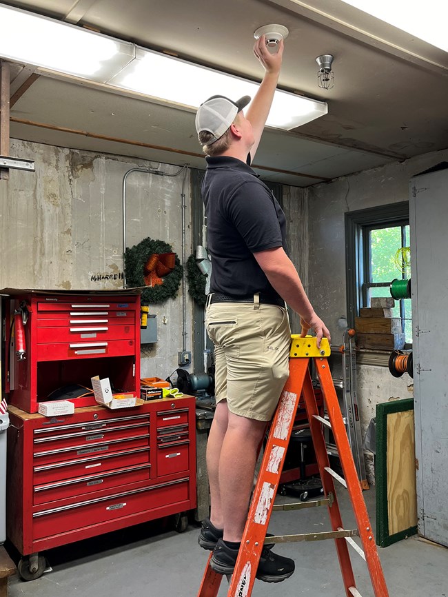 Holsenbeck, Ethan_FB_2_Photo A person stands on a ladder to reach a smoke detector on the ceiling.