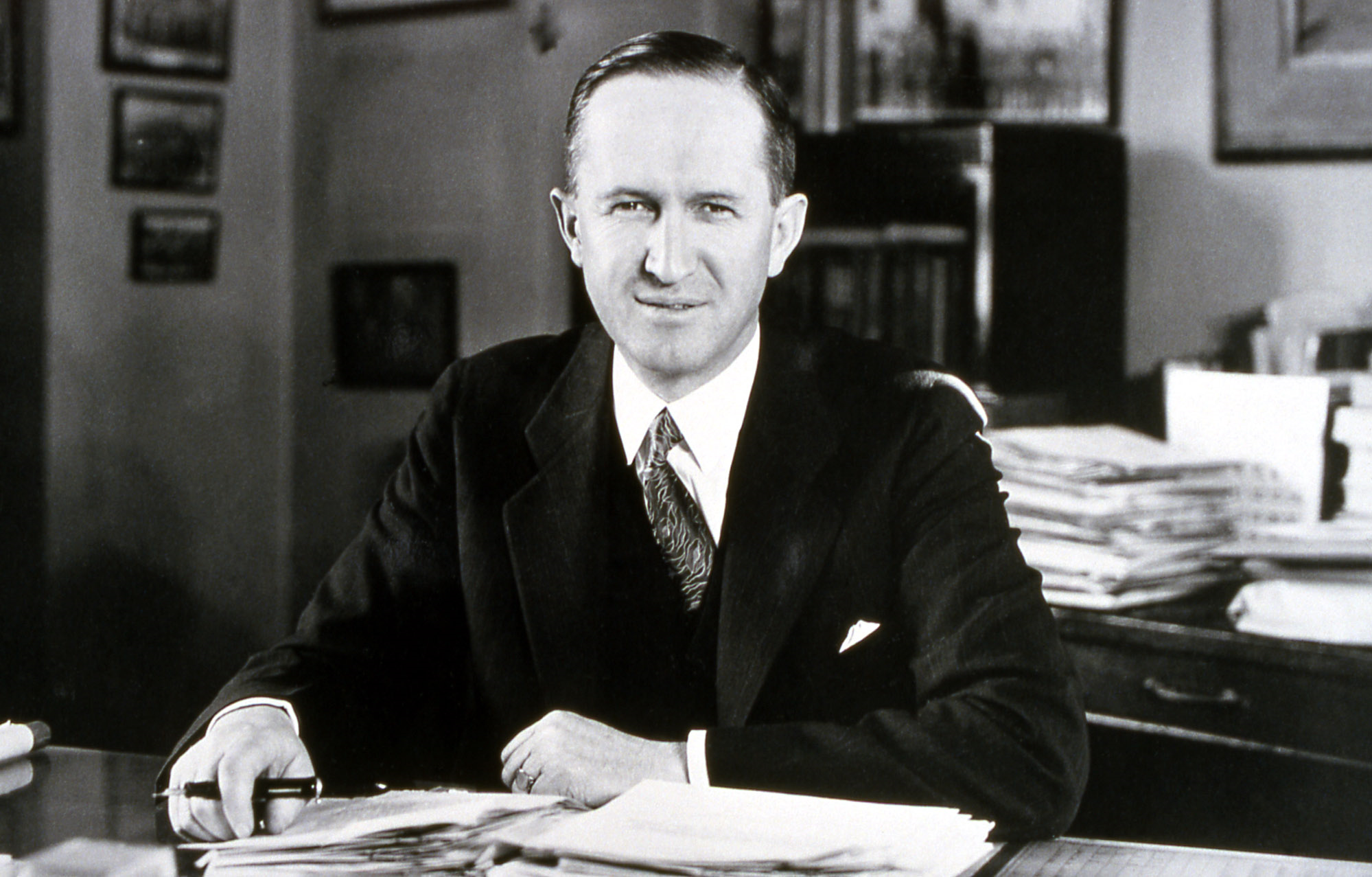 B&W photo of a man wearing a black suit sitting at a desk.