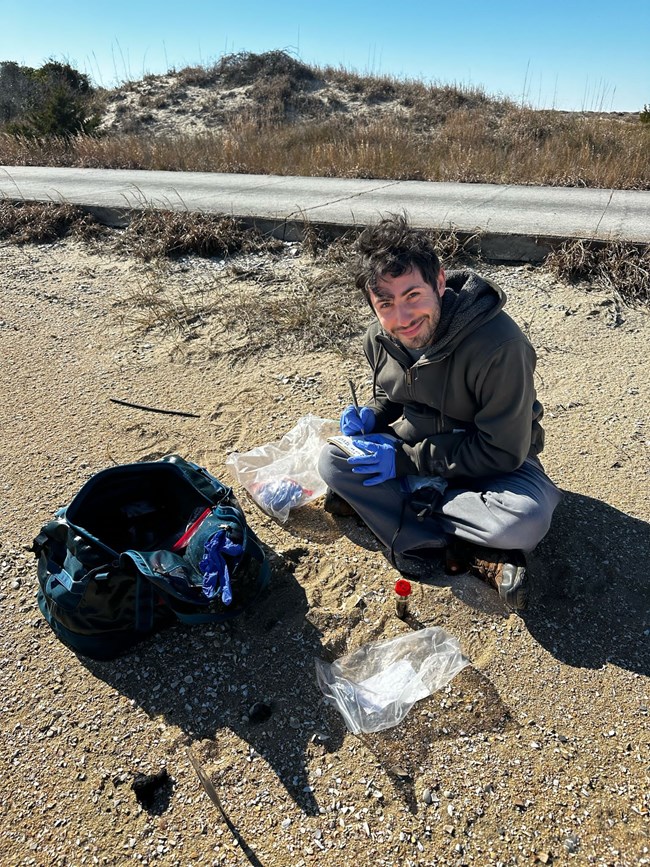 A person sitting in sand next to a road. He is wearing gloves and has field gear spread out beside him.